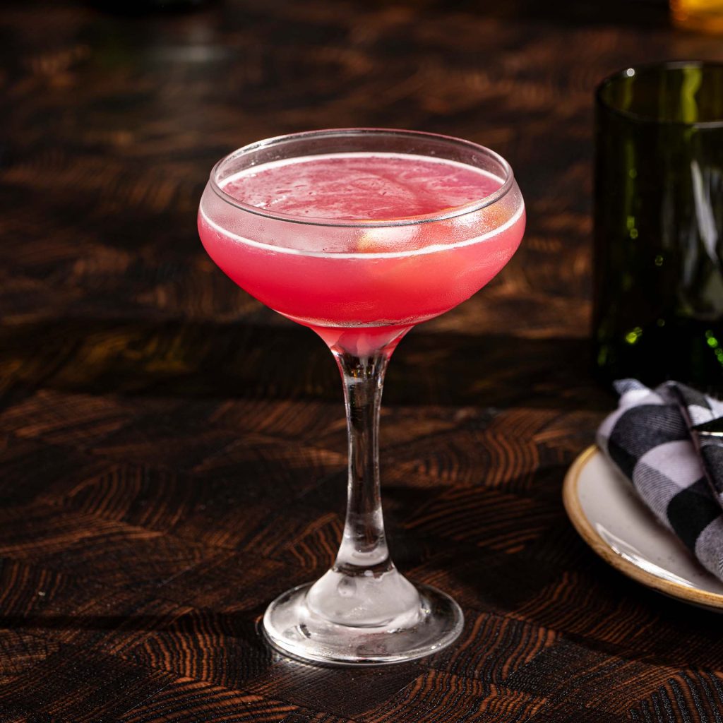 A cocktail in a stemmed glass sits on a wood table.